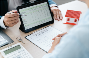 A professional points to financial data on a tablet while sitting at a desk with a contract, house model, and calculator.