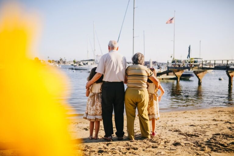An older couple stands on a beach with their arms around two young girls, all looking out toward sailboats in a harbor.